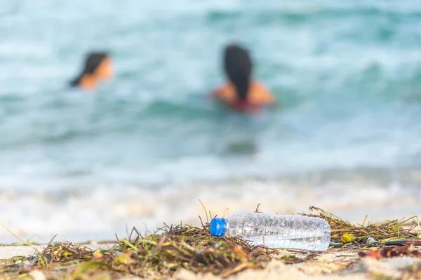 Close up image of empty plastic water bottle on dirty beach filled with seaweed, garbage and waste on dirty sandy beach with people in sea on background — Stock Photo, Image