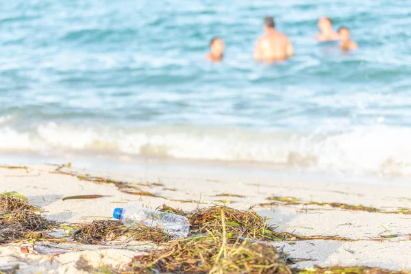 Yosun, çöp ve arka planda denizde insanlar ile kirli kumlu plajda atık dolu kirli plajda boş plastik su şişesi yakın görüntü — Stok fotoğraf