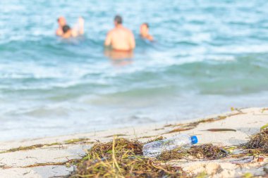 yosun, çöp ve arka planda denizde insanlar ile kirli kumlu plajda atık dolu kirli plajda boş plastik su şişesi yakın görüntü