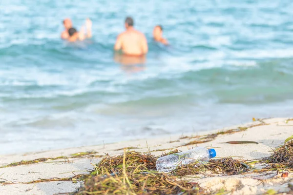 Close up image of empty plastic water bottle on dirty beach filled with seaweed, garbage and waste on dirty sandy beach with people in sea on background — Stock Photo, Image