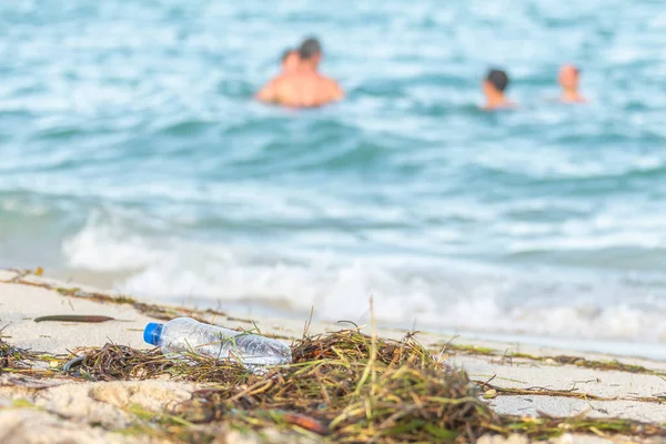 Immagine ravvicinata di bottiglia d'acqua di plastica vuota sulla spiaggia sporca piena di alghe, spazzatura e rifiuti sulla spiaggia di sabbia sporca con persone in mare sullo sfondo — Foto Stock