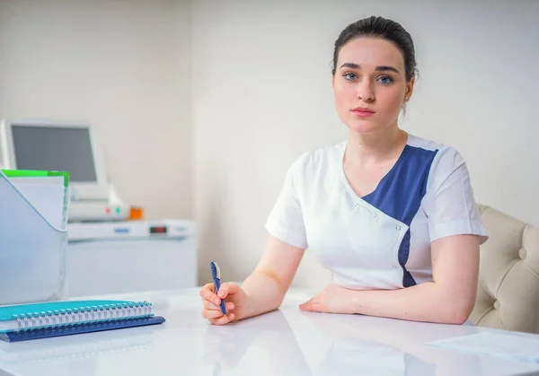 Masasında oturan genç bayan doktor elinde kalemle kameraya bakıyor. — Stok fotoğraf