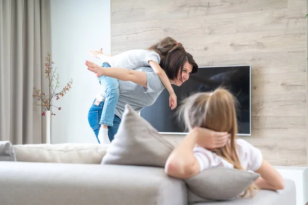 Preteen sad girl sitting on couch, während mutter having fun with younger sister, eifersucht concept — Stockfoto