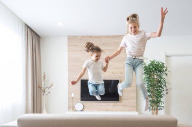 İki kız kardeş oturma odasında kanepede zıplayıp eğleniyorlar.