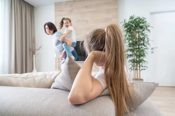 Preteen sad girl sitting on couch while mother having fun with younger sister, jealousy concept — Stock Photo, Image