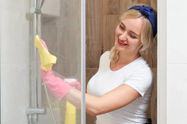 Jonge vrouw schoonmaken glazen douchecabine met spons en spuitfles — Stockfoto