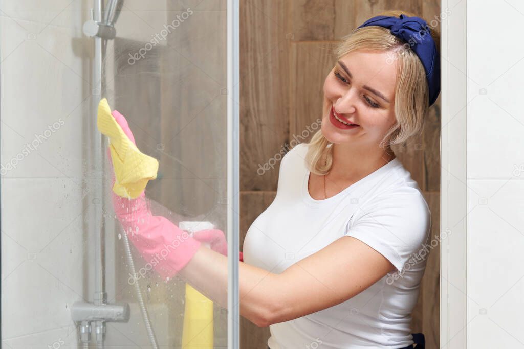 Young woman cleaning glass shower cabin with sponge and spray bottle