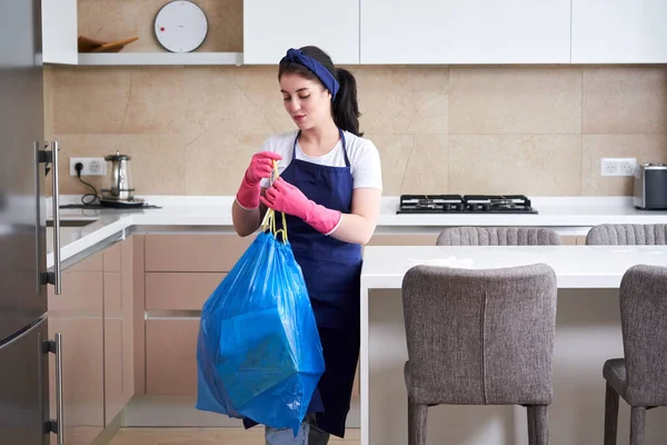 Домогосподарка в захисних гумових рукавичках тримає мішок для сміття, що стоїть на кухні. Концепція клінінгової служби — стокове фото