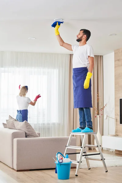 Man cleaning ceiling and lamps in living room. House cleaning service concept. — Stock Photo, Image