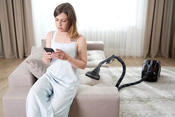 Mujer joven sentada en el sofá y usando el teléfono móvil mientras limpia en casa, copia el espacio. Concepto de tareas domésticas y tareas domésticas. Aparatos de limpieza —  Fotos de Stock