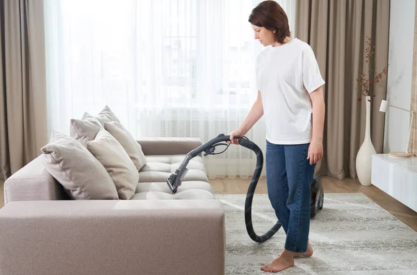 Junge Frau in weißem Hemd und Jeans putzt Teppich unter Sofa mit Staubsauger im Wohnzimmer, Kopierraum. Haushalts-, Reinigungs- und Hauswirtschaftskonzept — Stockfoto