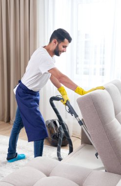 Evdeki elektrik süpürgesiyle kanepeyi temizleyen genç adam.