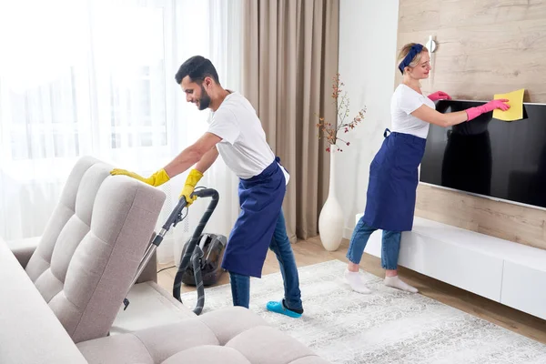 Mavi üniformalı profesyonel temizlikçiler dairenin oturma odasındaki mobilyaların tozunu siliyorlar. Temizlik hizmeti kavramı — Stok fotoğraf