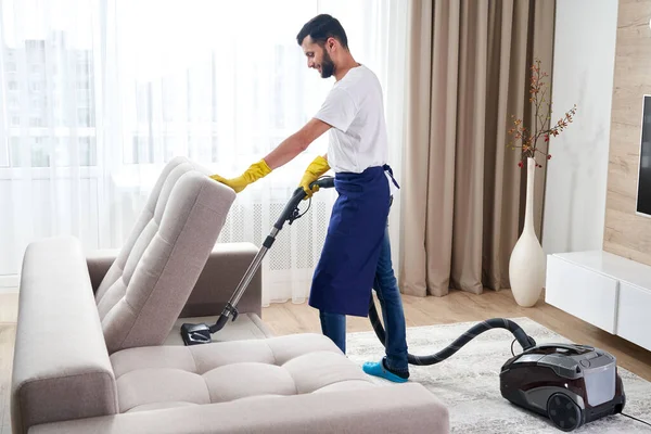 Молодой человек Уборка дивана с пылесосом очиститель в выходе из комнаты дома — стоковое фото