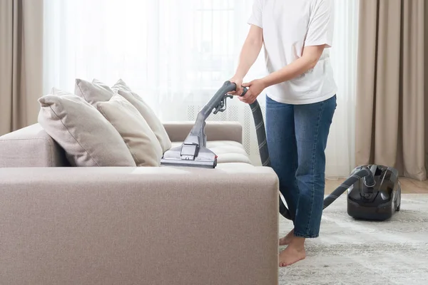 Młoda kobieta w białej koszuli i dżinsy czyszczenia dywanu pod kanapą z odkurzaczem w salonie, miejsca do kopiowania. Koncepcja prac domowych, sprzątania i prac domowych — Zdjęcie stockowe