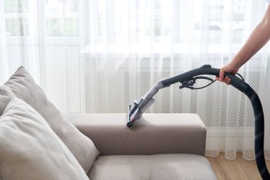 Genç ev hanımı oturma odasında elektrikli süpürgeyle kanepeyi temizliyor.