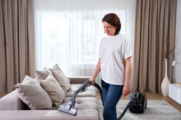 Молода жінка в білій сорочці і джинсах прибирає килим під диваном з пилососом у вітальні, копіює простір. Концепція домашнього завдання, чистки та клопоту — стокове фото