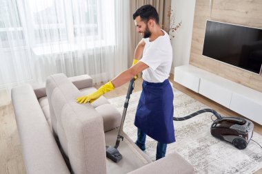 Evdeki elektrik süpürgesiyle kanepeyi temizleyen genç adam.
