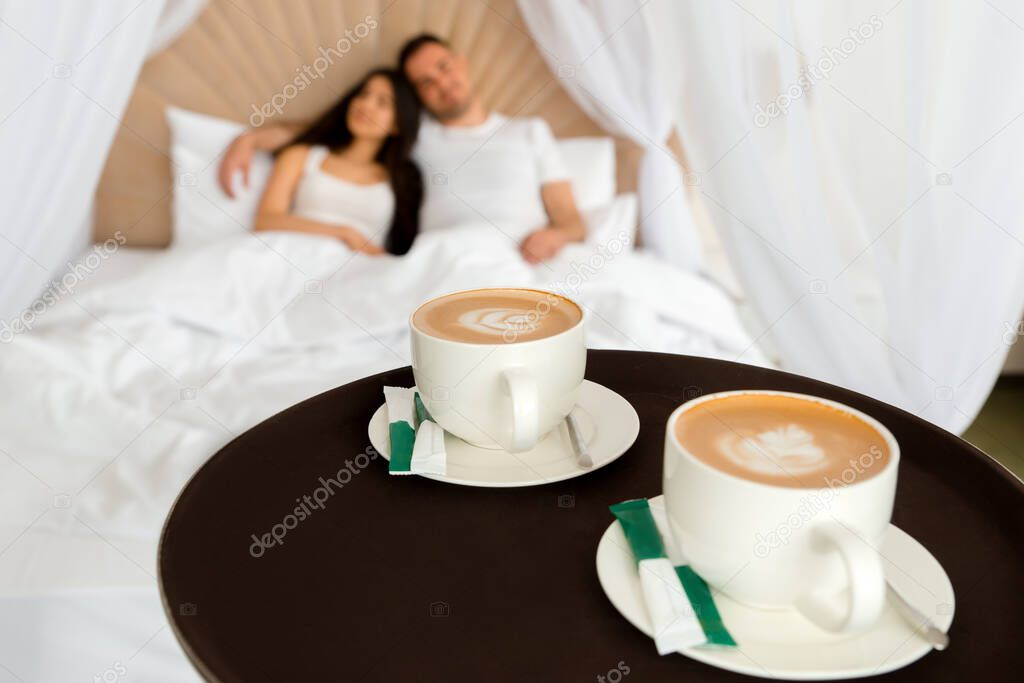 Room service delivering 2 cups of coffee to a hotel room for married couple lying in a bed in the morning