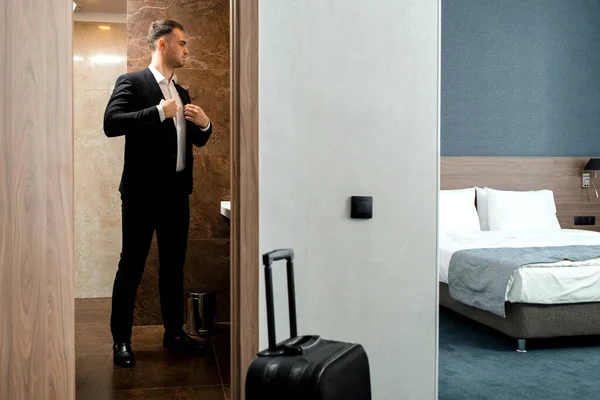 Mladý cestovatel podnikatel v bílé košili stojící u zrcadla a oblékání na setkání nebo práci po příjezdu do hotelového pokoje se zavazadly — Stock fotografie