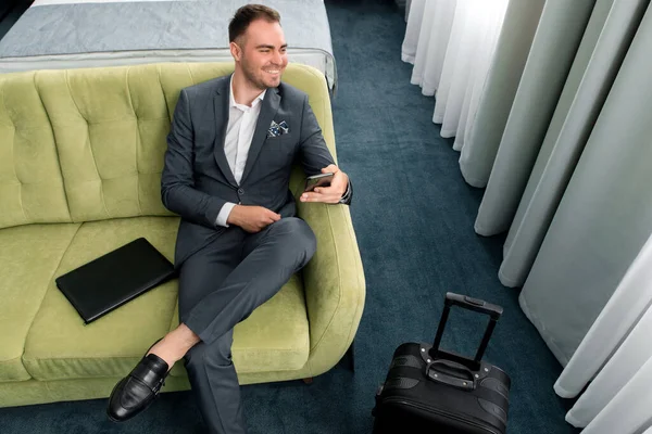 Fröhlicher Geschäftsmann sitzt auf Geschäftsreise mit Koffer im Hotelzimmer — Stockfoto