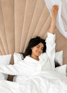 Yatakta kolları havada uzanan bir kadın. Çekici bir kızın portresi. Yorganın altında uzandıktan sonra yatakta eğleniyor.