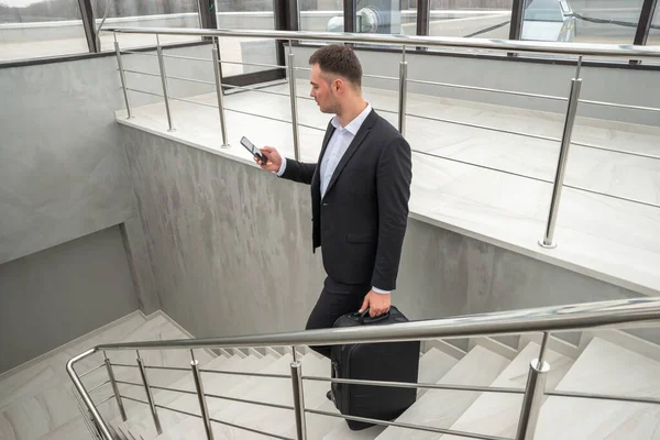 Joven hombre de negocios con maleta en traje elegante hablando por teléfono mientras camina por las escaleras en el moderno edificio de negocios — Foto de Stock