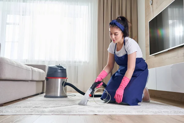 Женщина чистит ковер в гостиной, используя пылесос дома. Концепция услуги уборки — стоковое фото