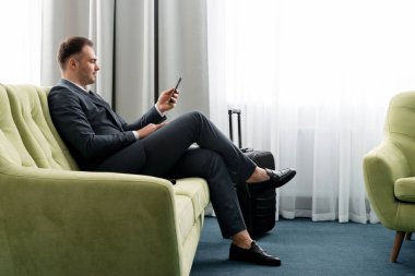 Neşeli iş adamı otel odasında bavuluyla iş gezisinde telefon kullanıyor.