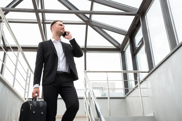 Joven hombre de negocios con maleta en traje elegante hablando por teléfono mientras camina por las escaleras en el moderno edificio de negocios — Foto de Stock