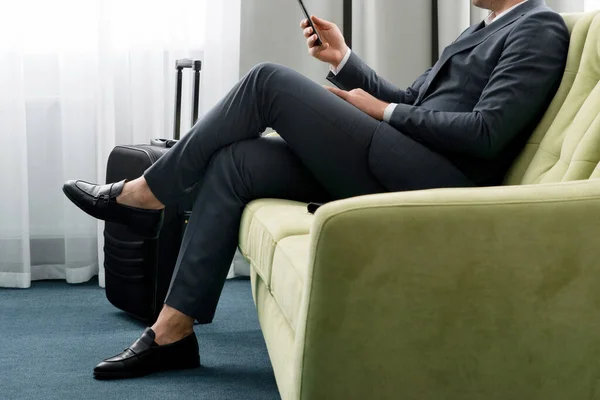 Hombre de negocios alegre usando teléfono sentado en la habitación del hotel con maleta en viaje de negocios — Foto de Stock