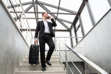 Modern iş binasında merdivenlerde yürürken şık takım elbiseli genç bir işadamı telefonla konuşuyor.