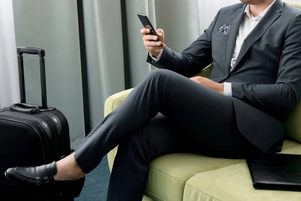Веселий бізнесмен використовує телефон, сидячи в готельному номері з валізою у відрядженні — стокове фото