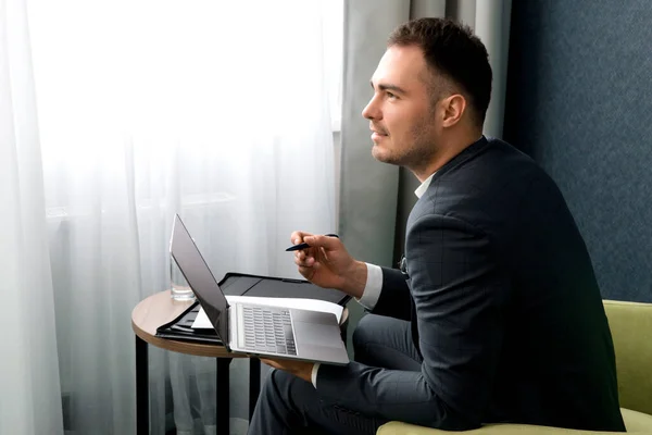 Joven hombre de negocios está utilizando el ordenador portátil mientras está sentado en la habitación del hotel con la maleta. — Foto de Stock