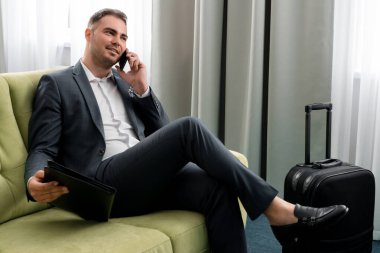 Neşeli iş adamı otel odasında bavuluyla iş gezisinde telefon kullanıyor.