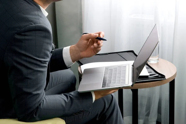 Junger Geschäftsmann arbeitet mit Laptop und Dokumenten, während er mit Koffer im Hotelzimmer sitzt. — Stockfoto