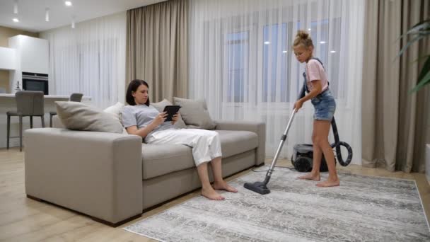 Reinigungskonzept. Junges Mädchen putzt Teppich mit Staubsauger, während Mutter Tablet benutzt — Stockvideo