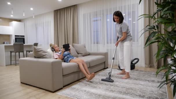 Piso de limpieza de la madre en la sala de estar con una aspiradora mientras su hija escucha música y el uso de la tableta mientras está sentado en el sofá — Vídeo de stock