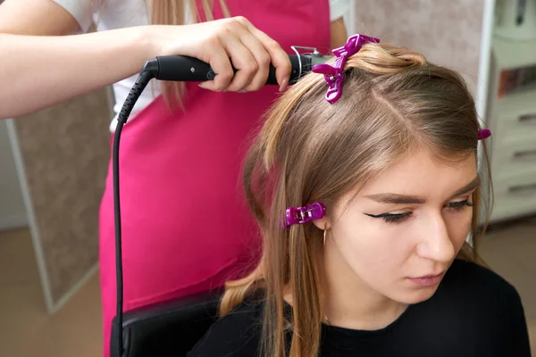 Cabeleireiro faz penteado menina com cabelo longo em um salão de beleza. Criar cachos com ferros de ondulação — Fotografia de Stock