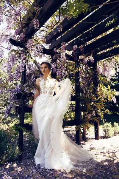时尚春天户外肖像美丽的年轻女子新娘在豪华婚纱和迪亚德马摆在盛开的紫藤树在阳光花园 — 图库照片