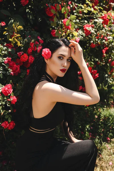 バラの茂みの花庭でポーズをとってエレガントな黒のドレスに黒髪の美しいセクシーな女性の屋外写真をファッションします — ストック写真