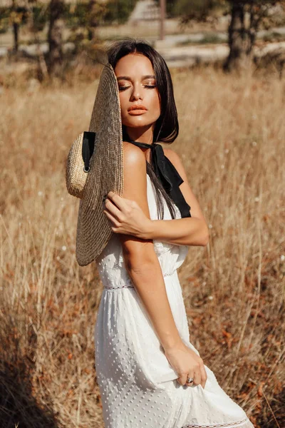 fashion summer outdoor photo of beautiful sexy woman with dark hair in elegant dress with luxurious straw hat and bag posing in field with dry grass