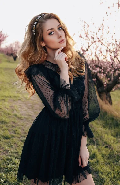 时尚户外照片的美丽感性的女孩与金发在优雅的黑色礼服和头带摆在盛开的桃花园 — 图库照片