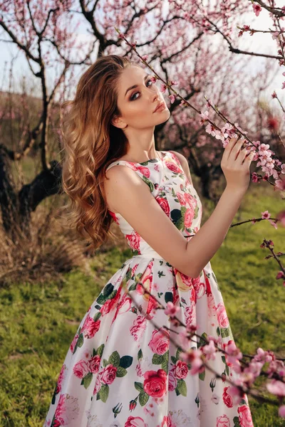 时尚户外照片的美丽感性的女孩与金发在优雅的礼服与花卉印花摆在盛开的桃花花园 — 图库照片