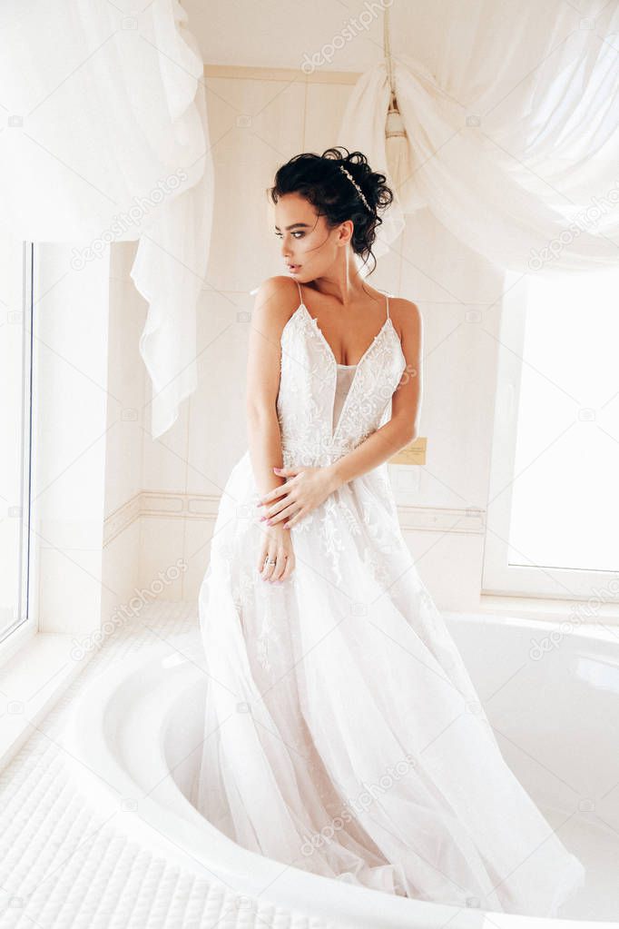 fashion interior photo of beautiful woman bride with dark hair in luxurious wedding dress posing in bath in bathroom