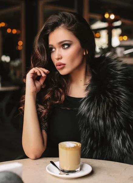 时尚照片美丽的年轻女子与黑头发在优雅的礼服坐在咖啡馆 桌上一杯卡布奇诺 — 图库照片