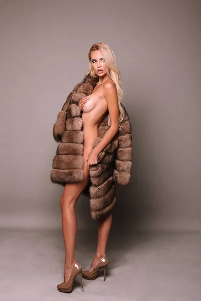 fashion photo of beautiful sexy woman with long blond hair in luxurious fur coat wearing on naked body  posing in studio