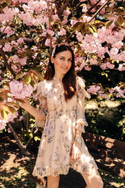 Koyu saçlı, zarif elbiseli, saku çiçeğinin yanında poz veren bir kadın.