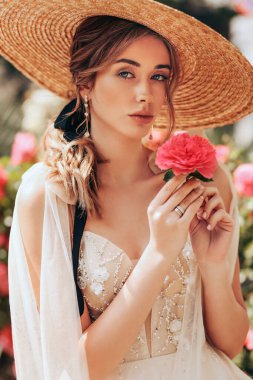 Havalı bir gelinlik içinde sarı saçlı güzel bir gelinin moda fotoğrafı ve bahar bahçesinde çiçek açan güllerle poz veren hasır bir şapka.