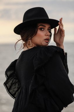 Güzel, siyah saçlı, şık elbiseli ve siyah şapkalı bir kadının plajda poz verdiği bir moda fotoğrafı.